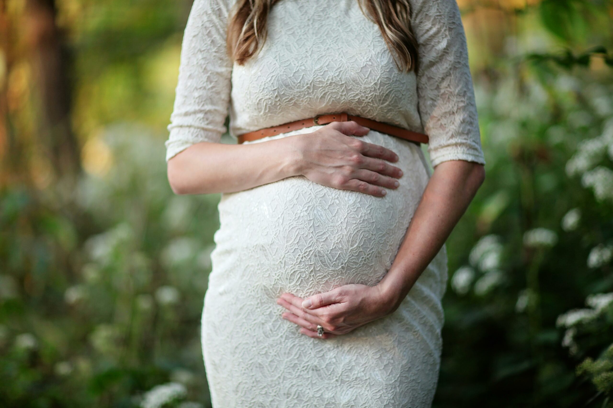 Zu sehen ist eine schwangere Frau in einem weißen Strickkleid, die ihren Bauch anfasst